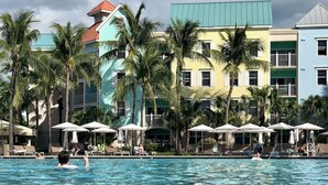 Une piscine où l’on voit des personnes dans l’eau, entourée de bâtiments aux couleurs vives et de palmiers.