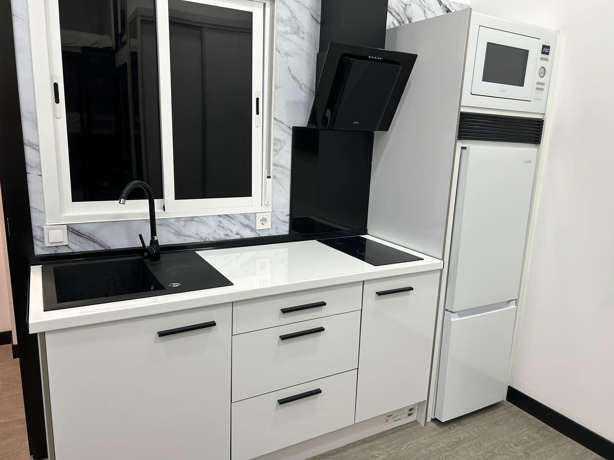 Cocina compacta con encimera blanca, grifo negro y microondas integrado.