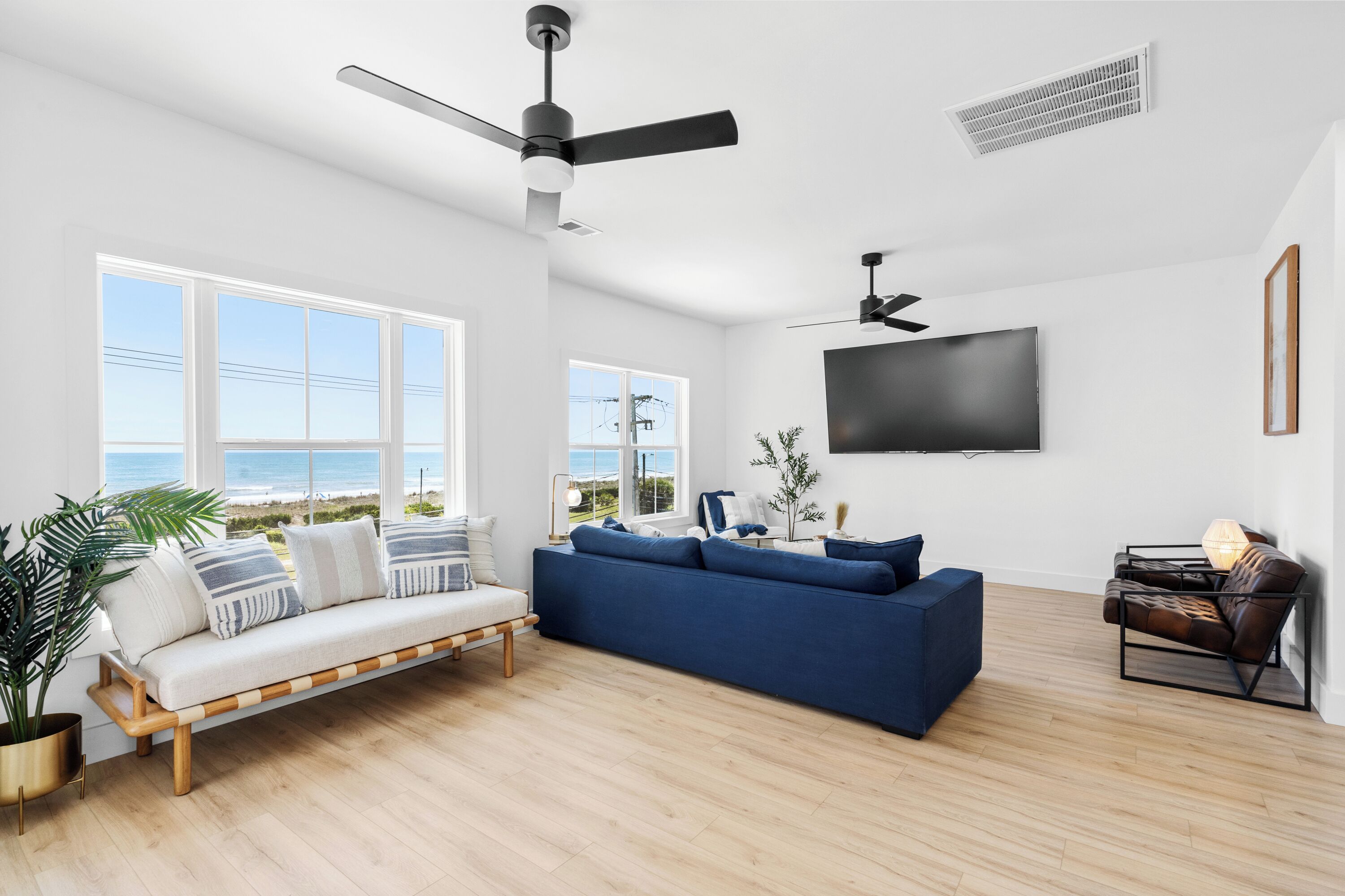 A modern living room with a blue sofa, a wooden bench, a TV mounted on the wall, and large windows with a view of the ocean.