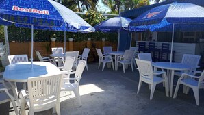 Un espace extérieur aménagé en terrasse, avec des chaises et des tables en plastique blanc, protégé par des parasols bleus arborant la marque « BestBull ».