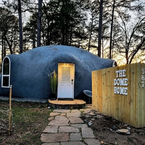 Eine kuppelförmige Struktur mit einer Tür und einem Holzzaun, auf dem 'THE DOME HOME' steht.