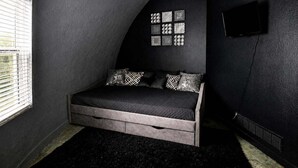 Une chambre à coucher à l’ambiance sombre, avec un lit, une fenêtre, un téléviseur fixé au mur et un ventilateur de plafond au design original.