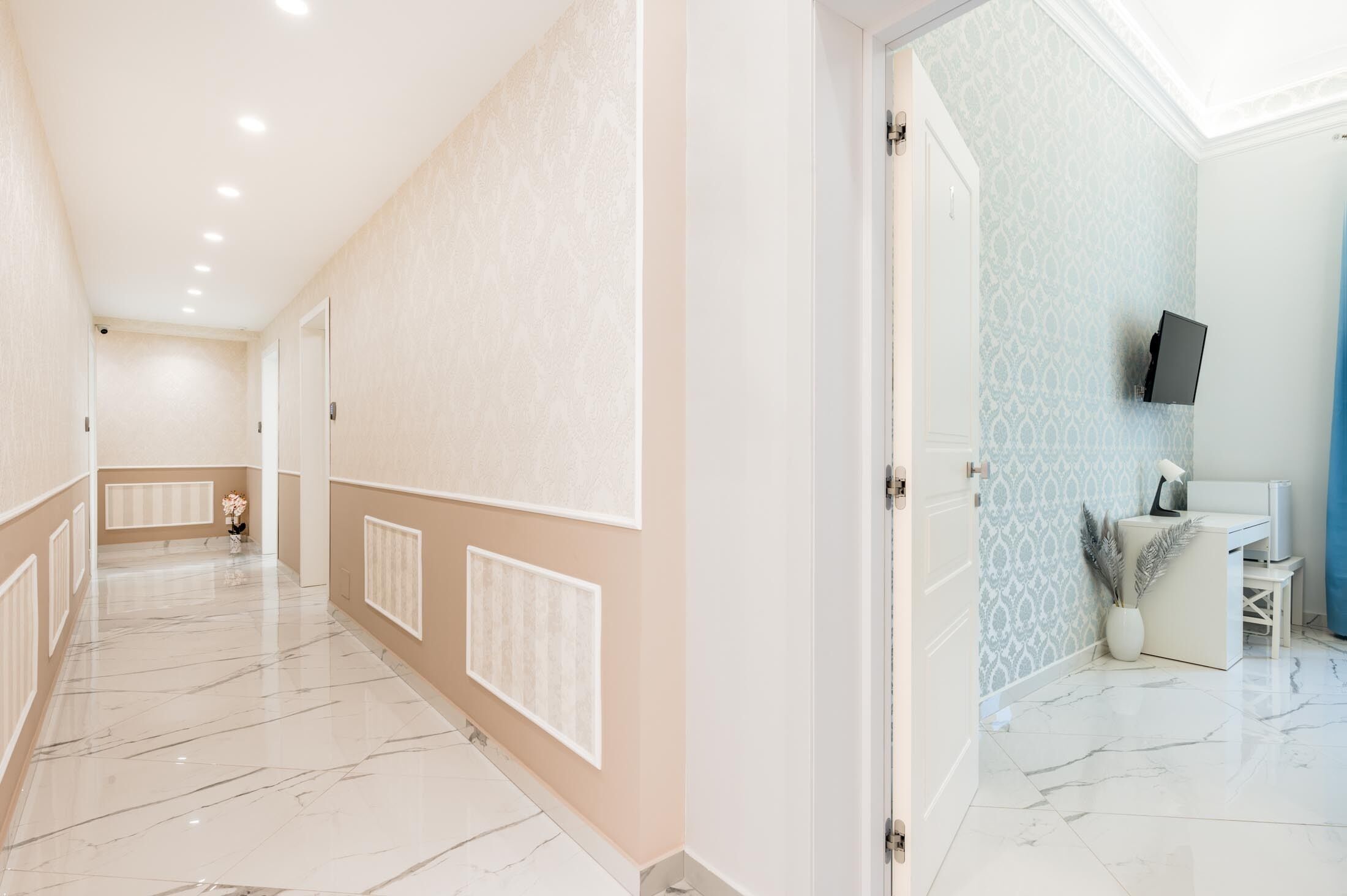 A hallway with a marble floor, beige walls, and a door leading to a room with a blue patterned wall and a mounted TV.