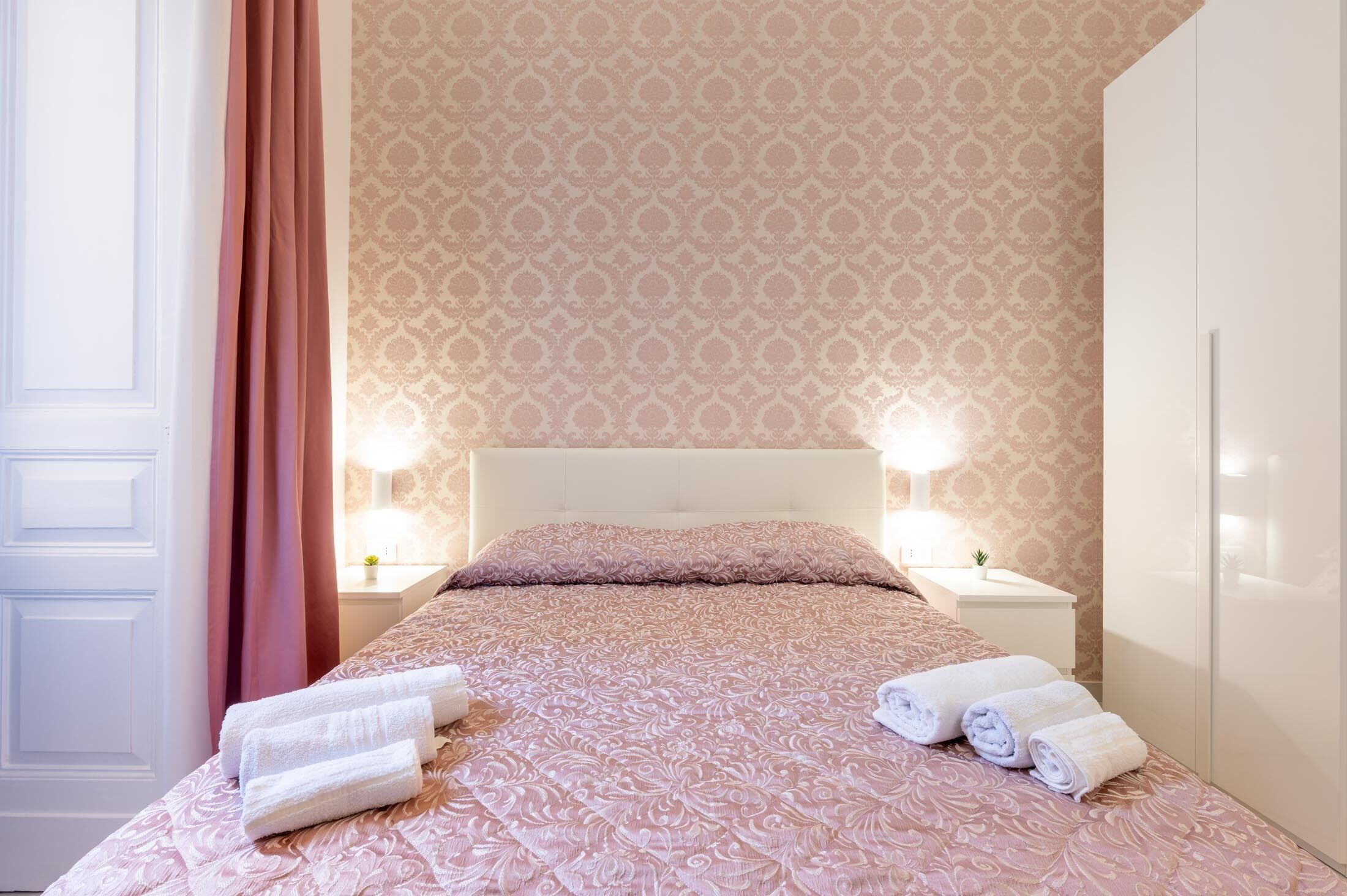 A neatly made bed with a patterned bedspread, two bedside tables with lamps, and a patterned wallpaper.