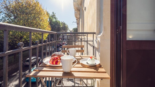 A balcony with a table set for breakfast, including croissants and fruit, overlooking a street with parked cars and trees. - Palazzo Bellini (Catania)