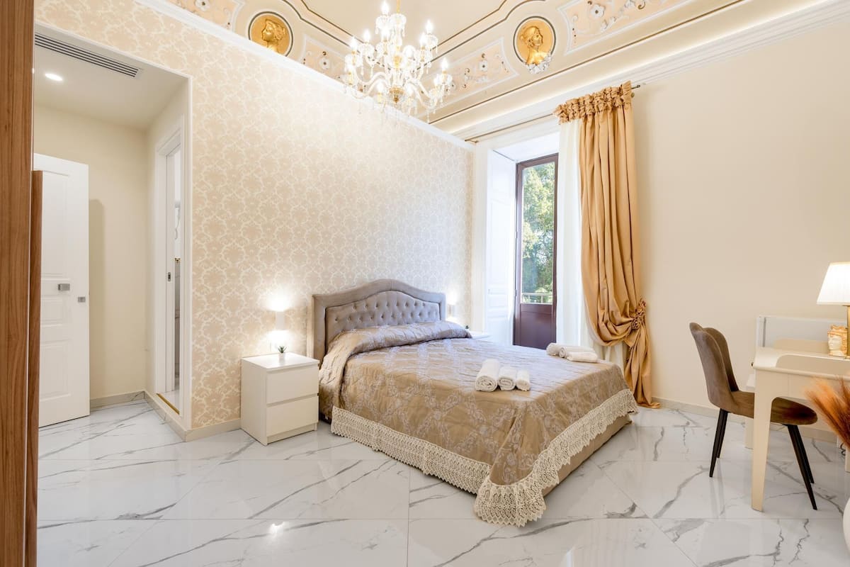 A bedroom with a large bed, a nightstand, a chair, and a desk with a lamp.