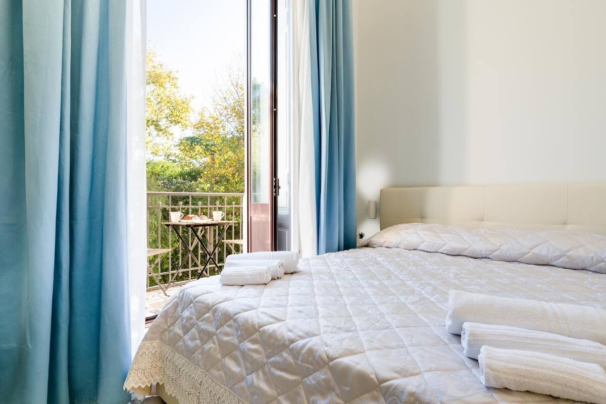 A bedroom with a large bed, white bedding, and a view of a balcony with a table and chairs.
