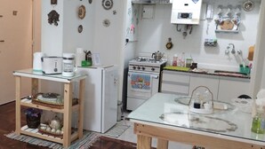 Una cocina con refrigerador, estufa y cafetera. Hay una alacena con vajilla y una mesita con cubierta de vidrio.