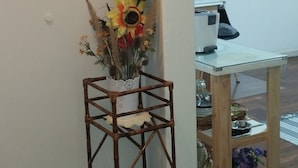 Un rincón de una habitación con una silla de madera, una mesa pequeña con un florero y un ventilador fijado a la pared.
