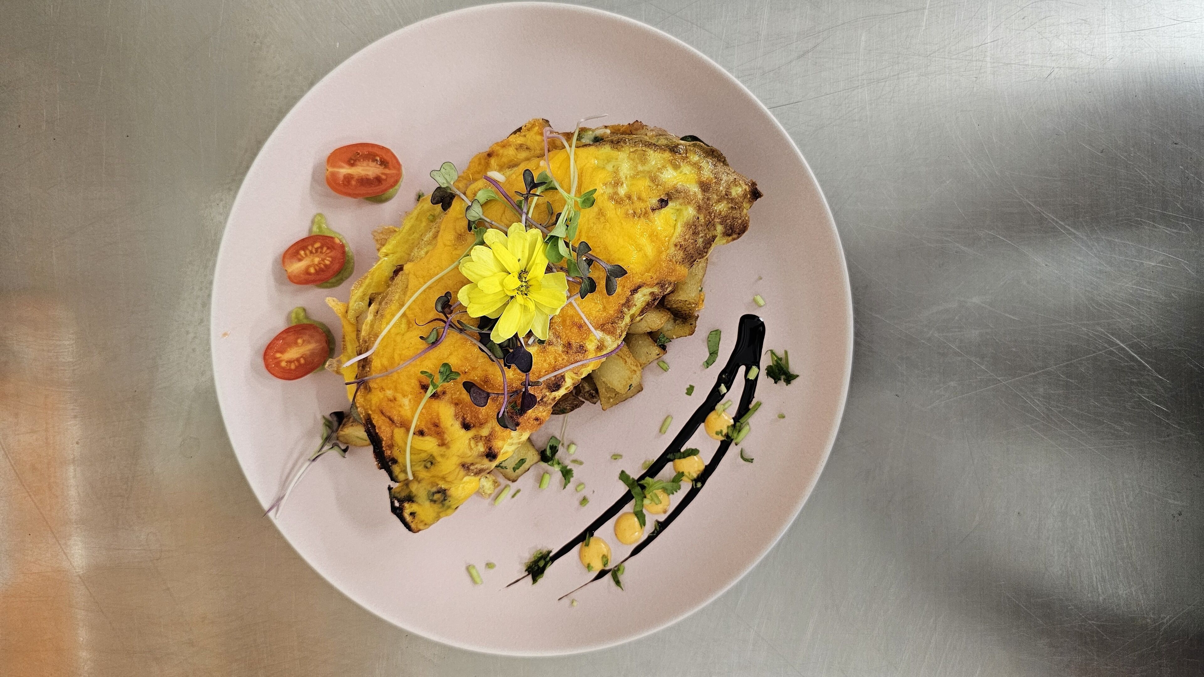 A plate with a grilled dish, garnished with herbs and a yellow flower, on a stainless steel surface.
