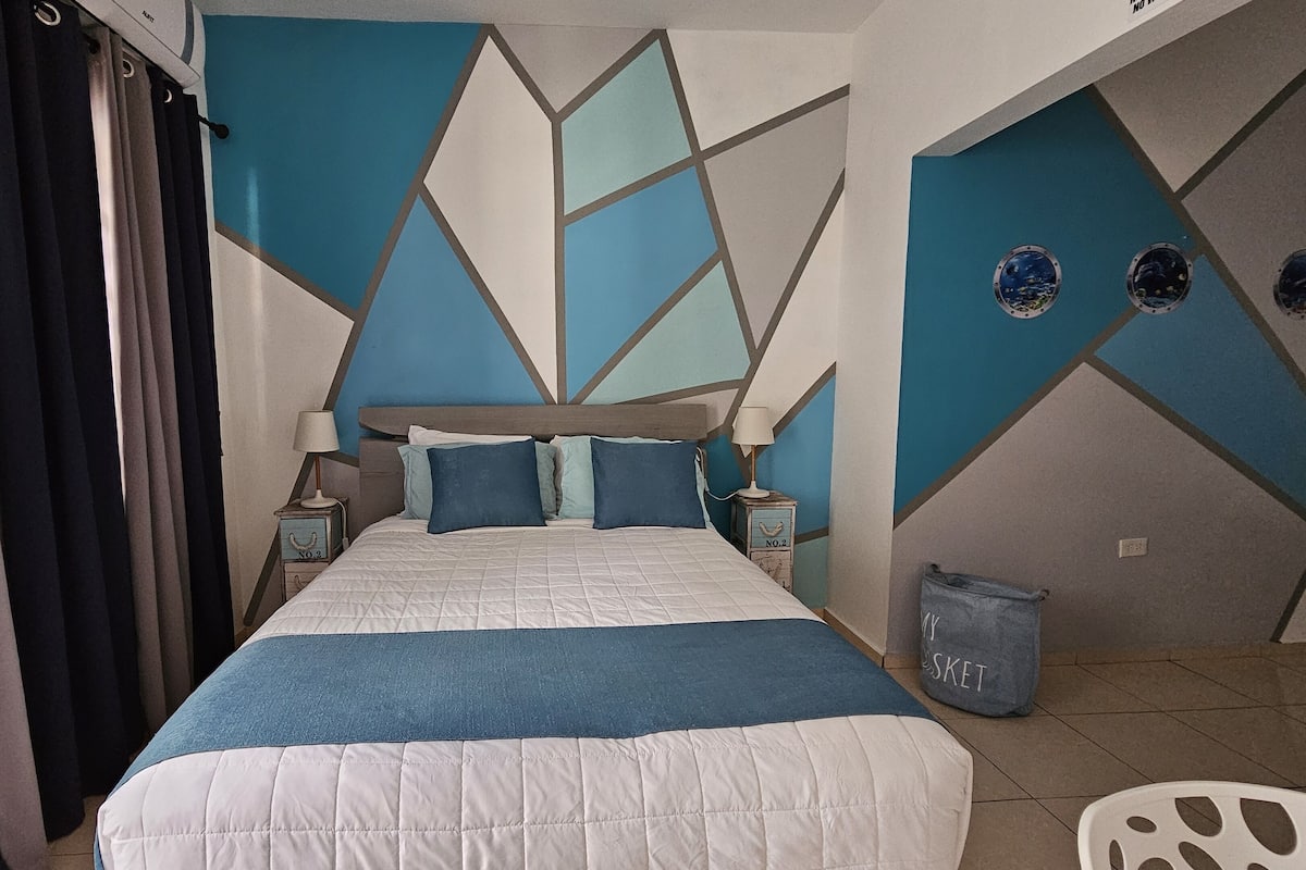 A bedroom with a bed, two bedside tables, a nightstand, a window with curtains, and a wall with geometric patterns.