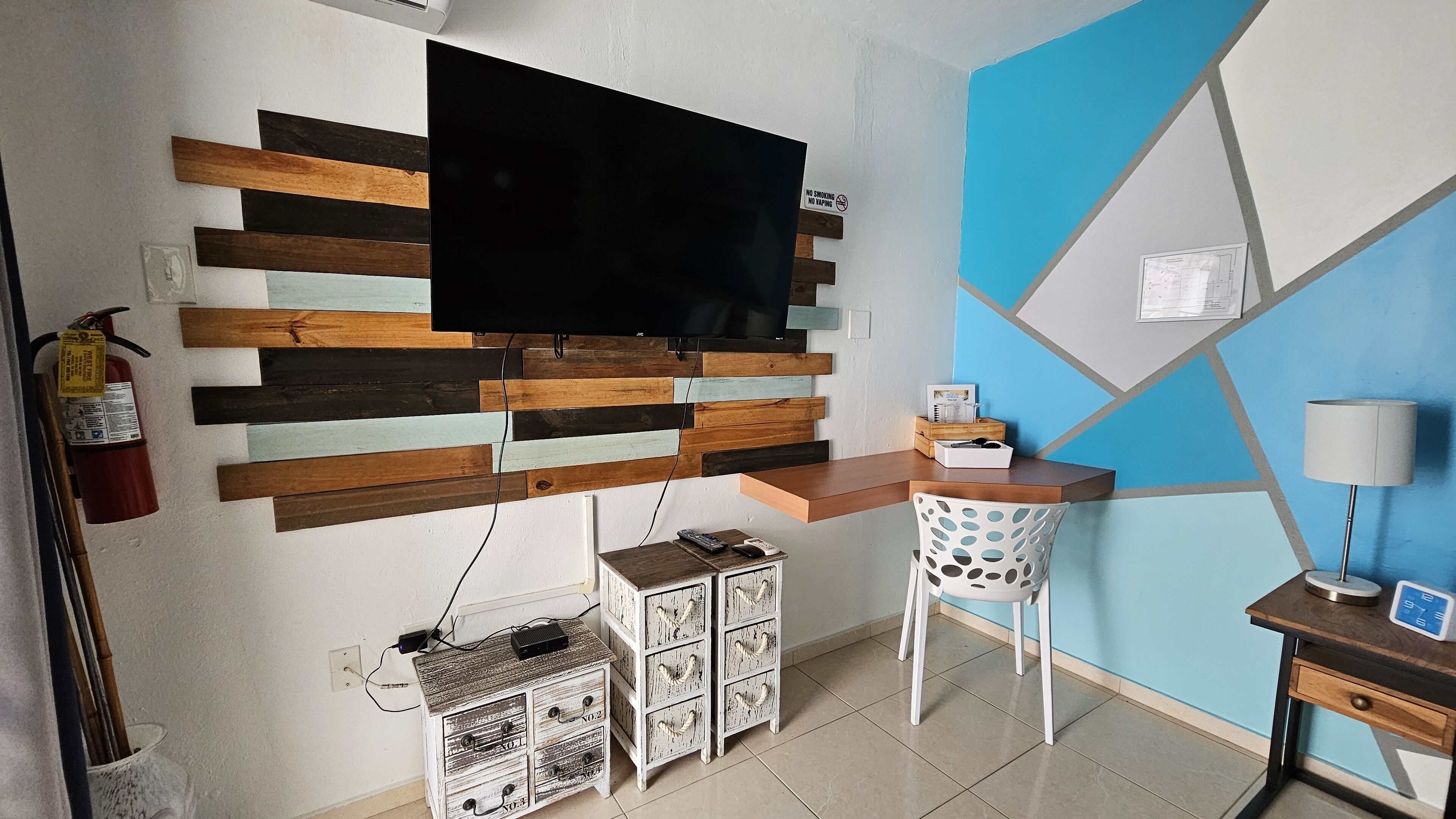 A modern living room with a flat-screen TV mounted on a wooden panel wall, a small wooden side table with a lamp, and a white chair.