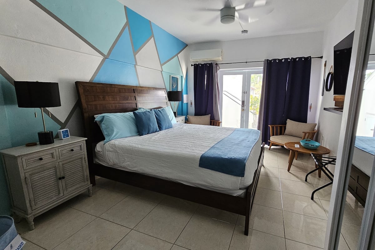 A bedroom with a bed, bedside table, lamp, and a view of the outdoors.