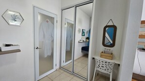 Habitación de hotel con puerta corrediza de vidrio, una bata blanca colgada en la puerta, un estante blanco con un jarrón y un espejo en la pared, además de un aparador de madera.