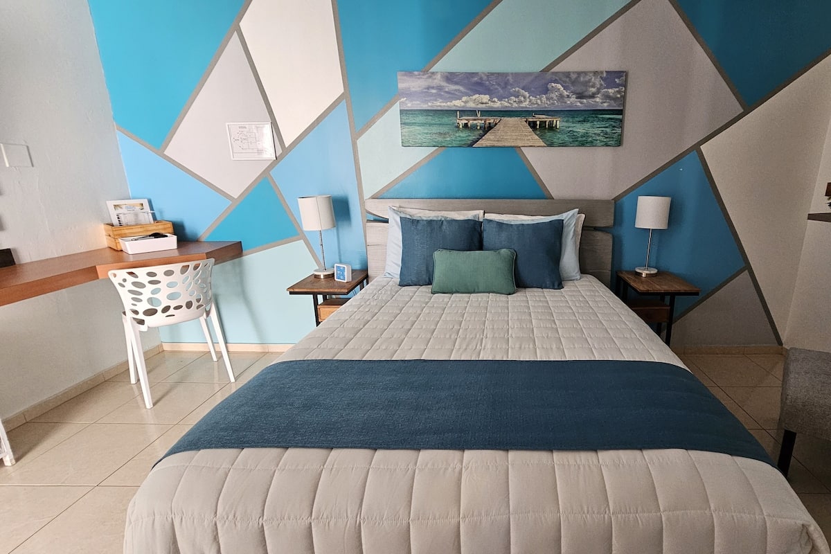 A modern hotel room with a geometric wall design, a bed with a blue and white duvet, two nightstands, a desk, and a chair.