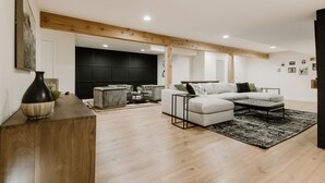 Un salon moderne avec un canapé d’angle, une table basse et un tapis. Le plafond est orné de poutres en bois et il y a un meuble de rangement noir pour les appareils électroniques.
