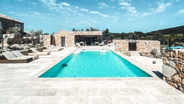 Una piscina con muros de piedra y sillones de descanso, rodeada de edificios de piedra y un cielo despejado.
