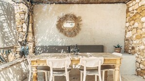 Un patio al aire libre con una mesa de madera y sillas de mimbre blanco, una pared de piedra y una guirnalda decorativa en la pared.