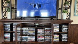 A home entertainment setup with a large flat-screen TV displaying a flight scene, surrounded by shelves of DVDs and Blu-rays.