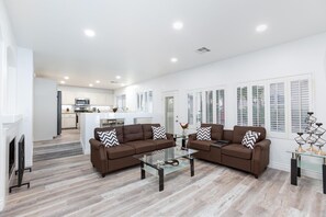 Una sala de estar moderna con dos sofás marrones, una mesa de centro de cristal y una cocina al fondo.