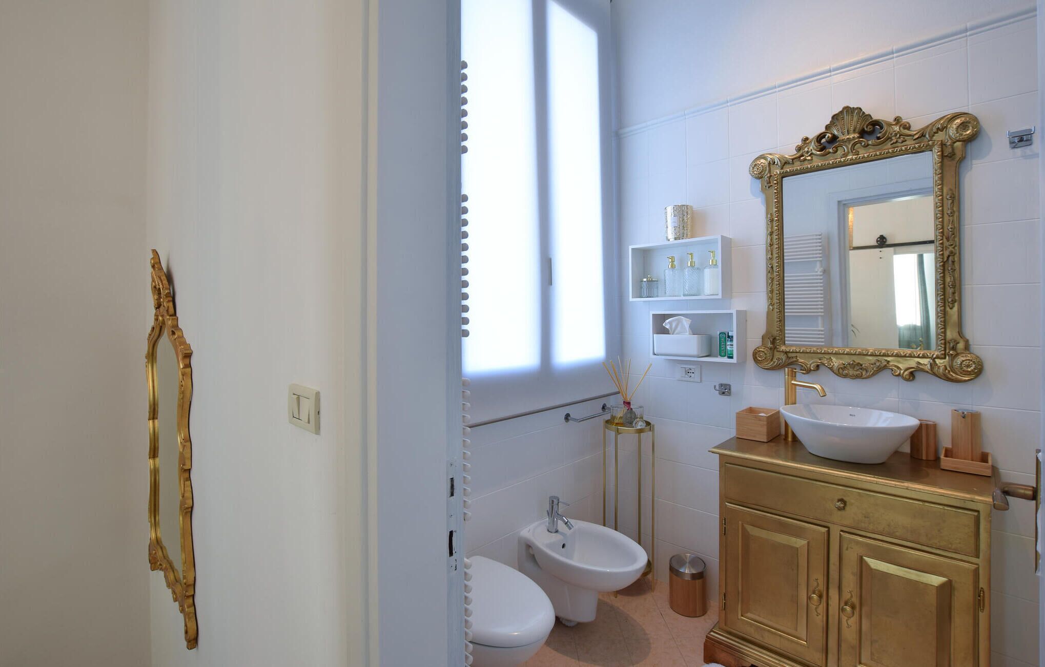 Un baño con inodoro blanco, un bidé, un lavabo con mueble dorado y un espejo grande y ornamentado.