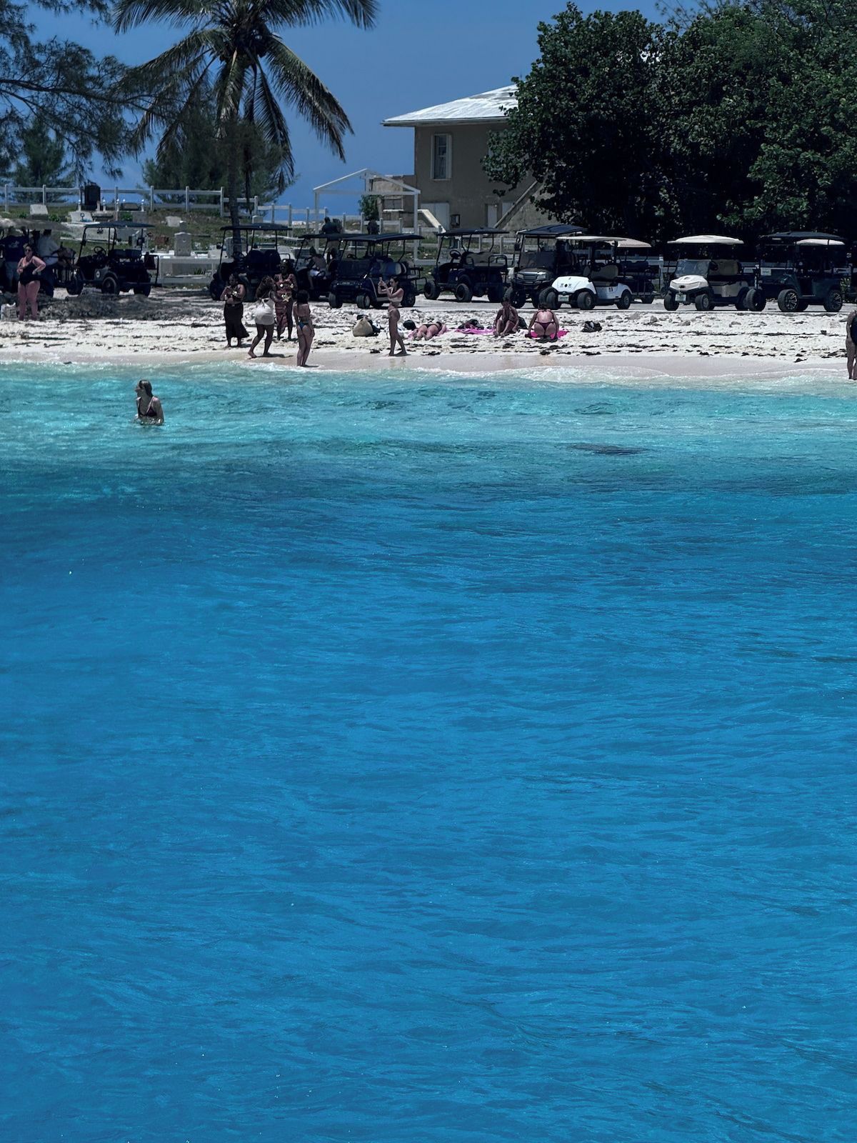 Ein Strand mit klarem, blauem Wasser, ein paar Personen und Golfcarts, die im Sand geparkt sind.