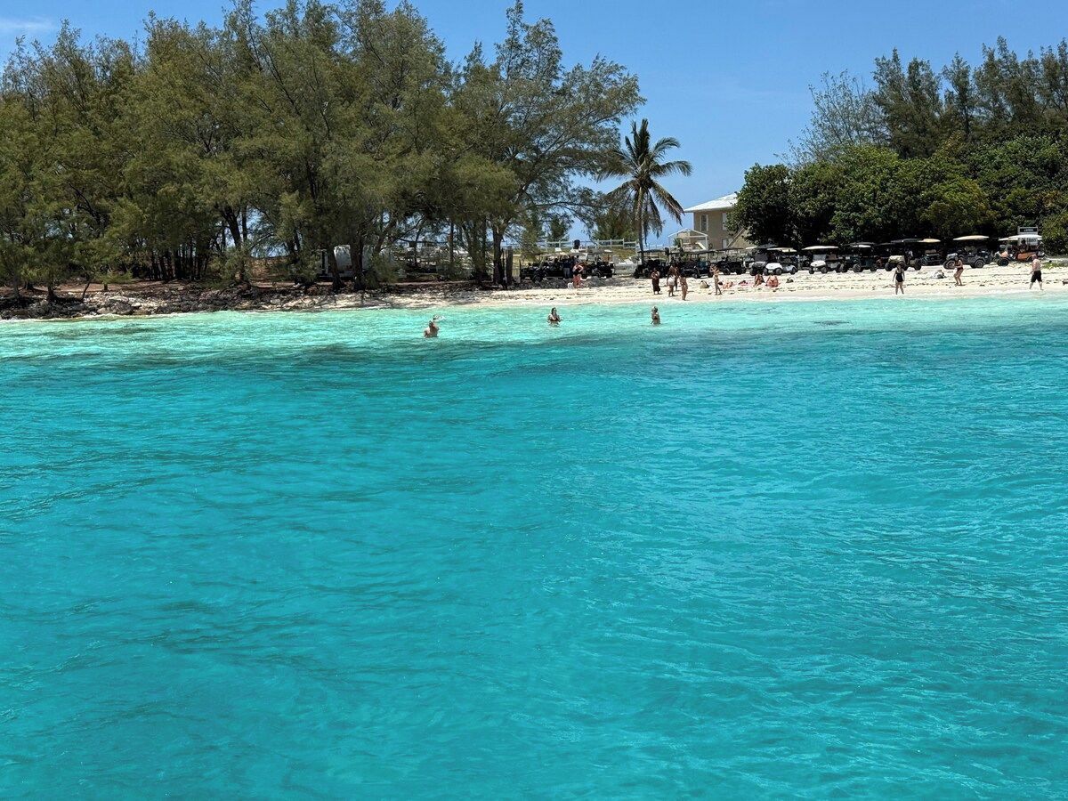 Une plage à l’eau turquoise et limpide, où l’on peut voir des gens se baigner, et une rangée d’arbres en arrière-plan.