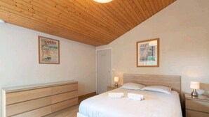 Een slaapkamer met een houten plafond, een bed met witte lakens, een nachtkastje met een lamp en een houten dressoir.