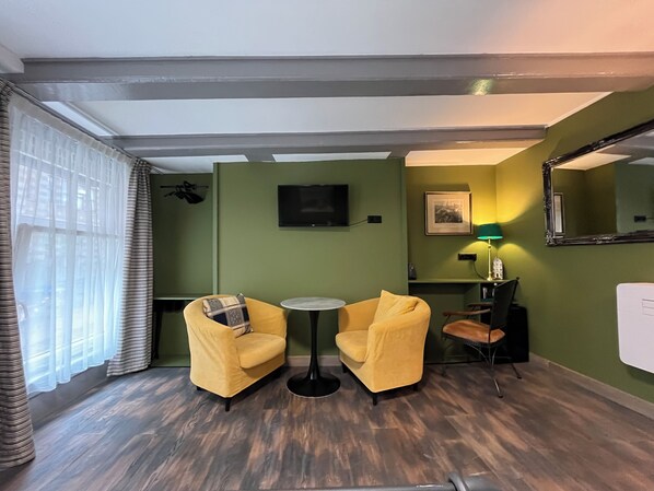 Una habitación con dos sillones amarillos, una pequeña mesa redonda, un televisor montado en la pared, un cuadro enmarcado, un espejo y una lámpara.