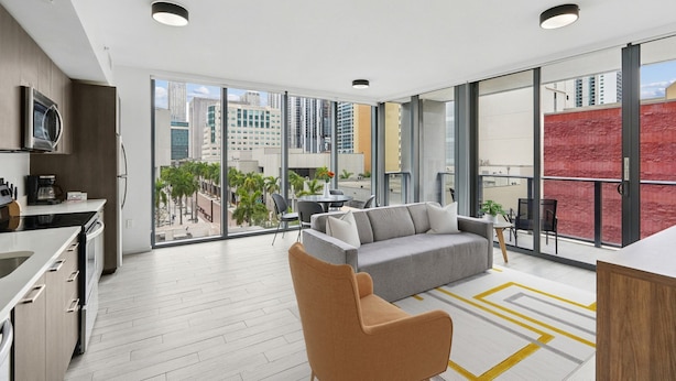 A modern apartment with a kitchen, a grey sofa, a brown armchair, and large windows offering a city view. - Sentral Alea Miami (Miami)
