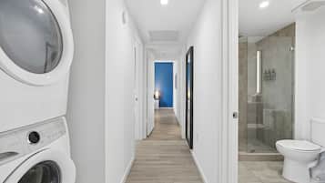 Une buanderie équipée d’un lave-linge et d’un sèche-linge, un couloir menant à une salle de bain avec une douche à parois de verre, et un mur d’accent bleu.