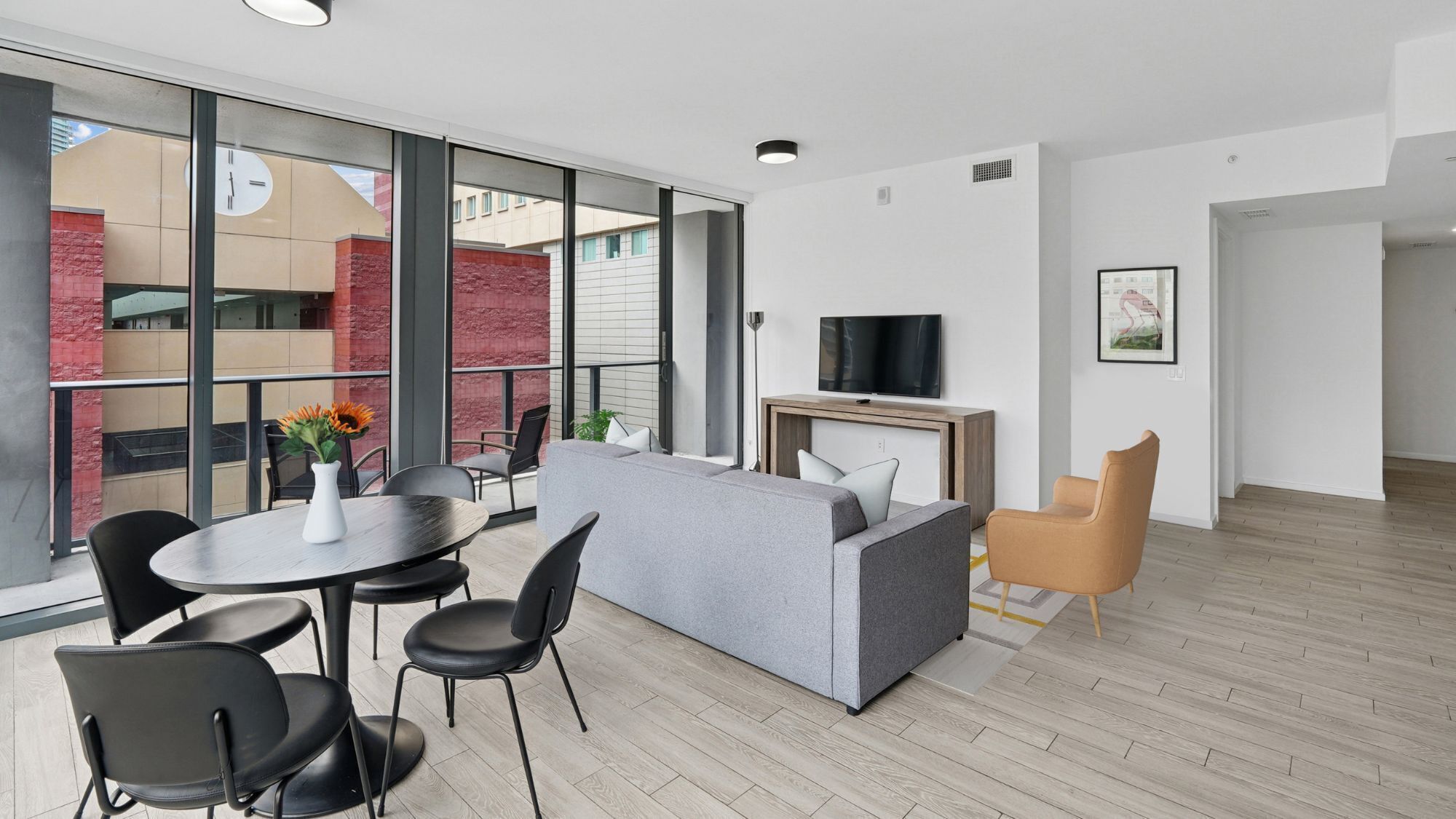 A modern living room with a grey sofa, black chairs, a round table with a vase, a flat-screen TV, and a wooden coffee table.