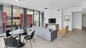A modern living room with a grey sofa, black chairs, a round table with a vase, a flat-screen TV, and a wooden coffee table.
