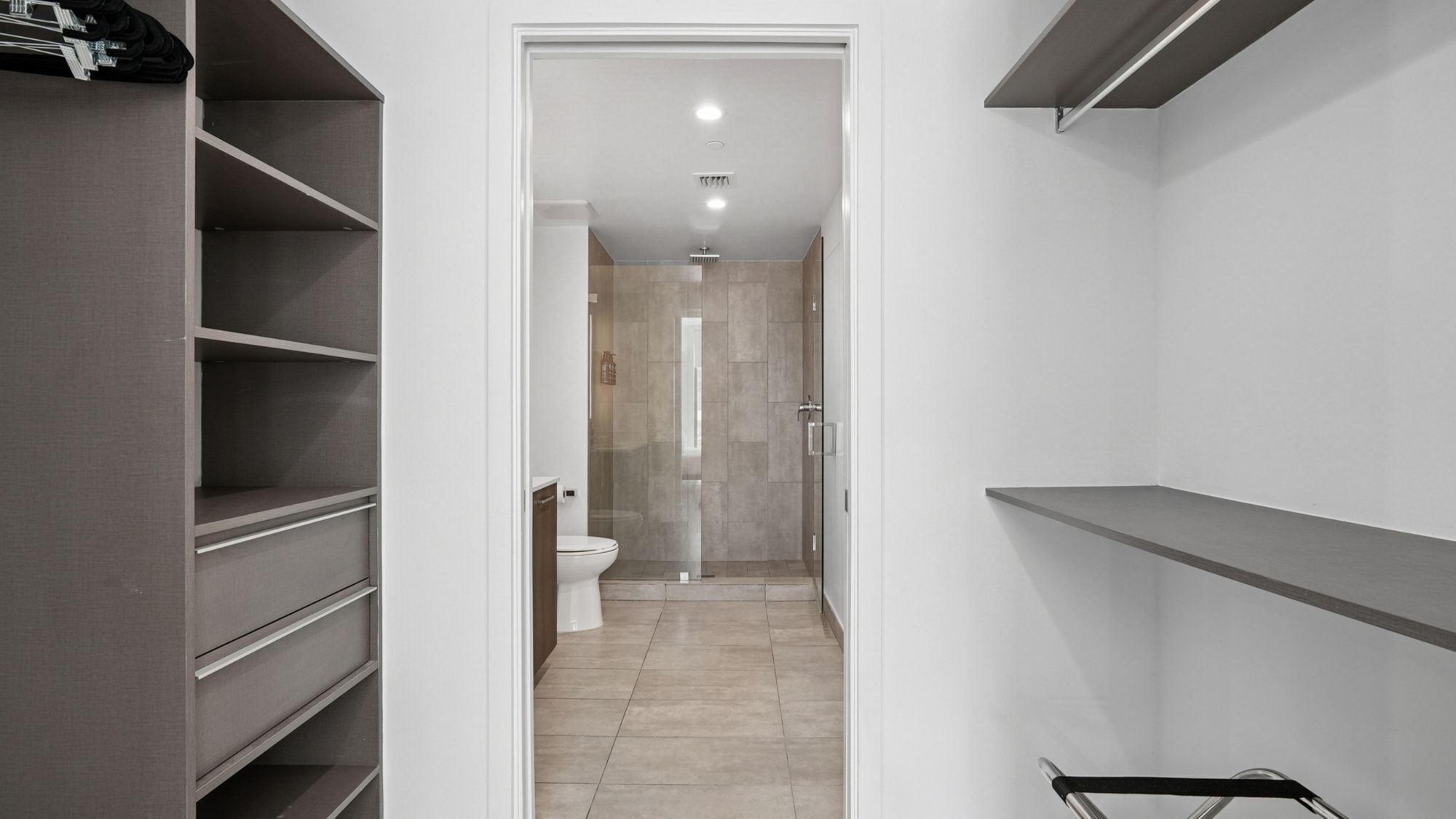 Un couloir étroit mène à une salle de bain équipée de toilettes et d’une douche. Des étagères sont disposées de part et d’autre du couloir.