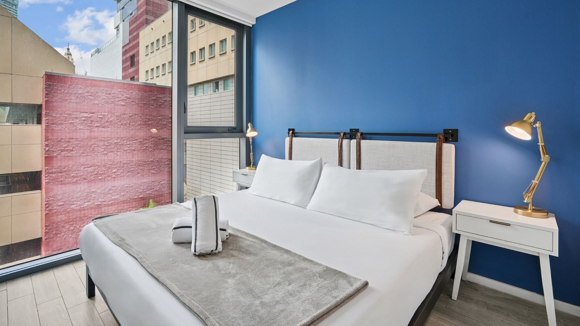 A modern hotel room with a large bed, a nightstand, a desk lamp, and a view of buildings through a large window.