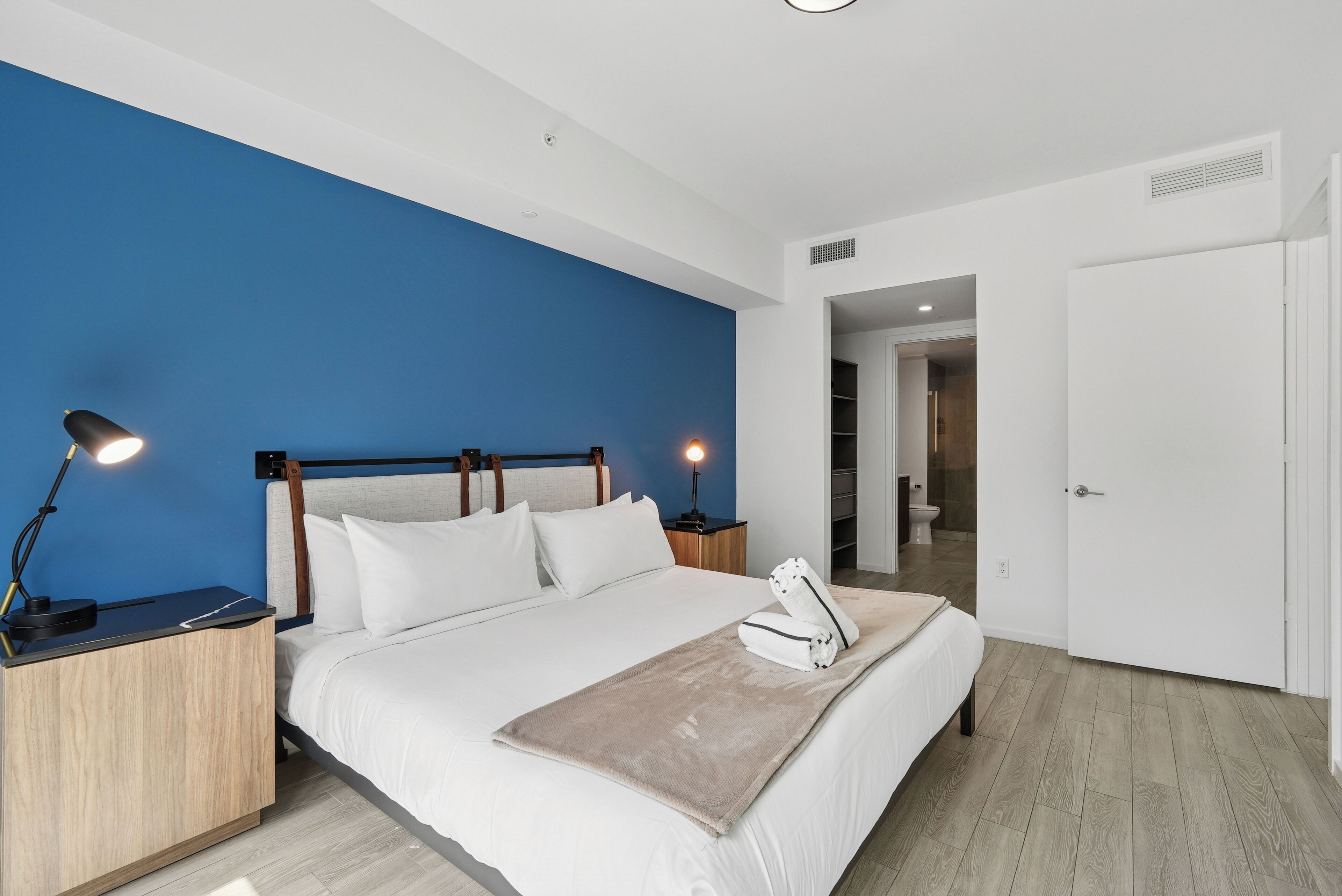 A modern bedroom with a large bed, two bedside lamps, a nightstand, and a bathroom visible through an open door.