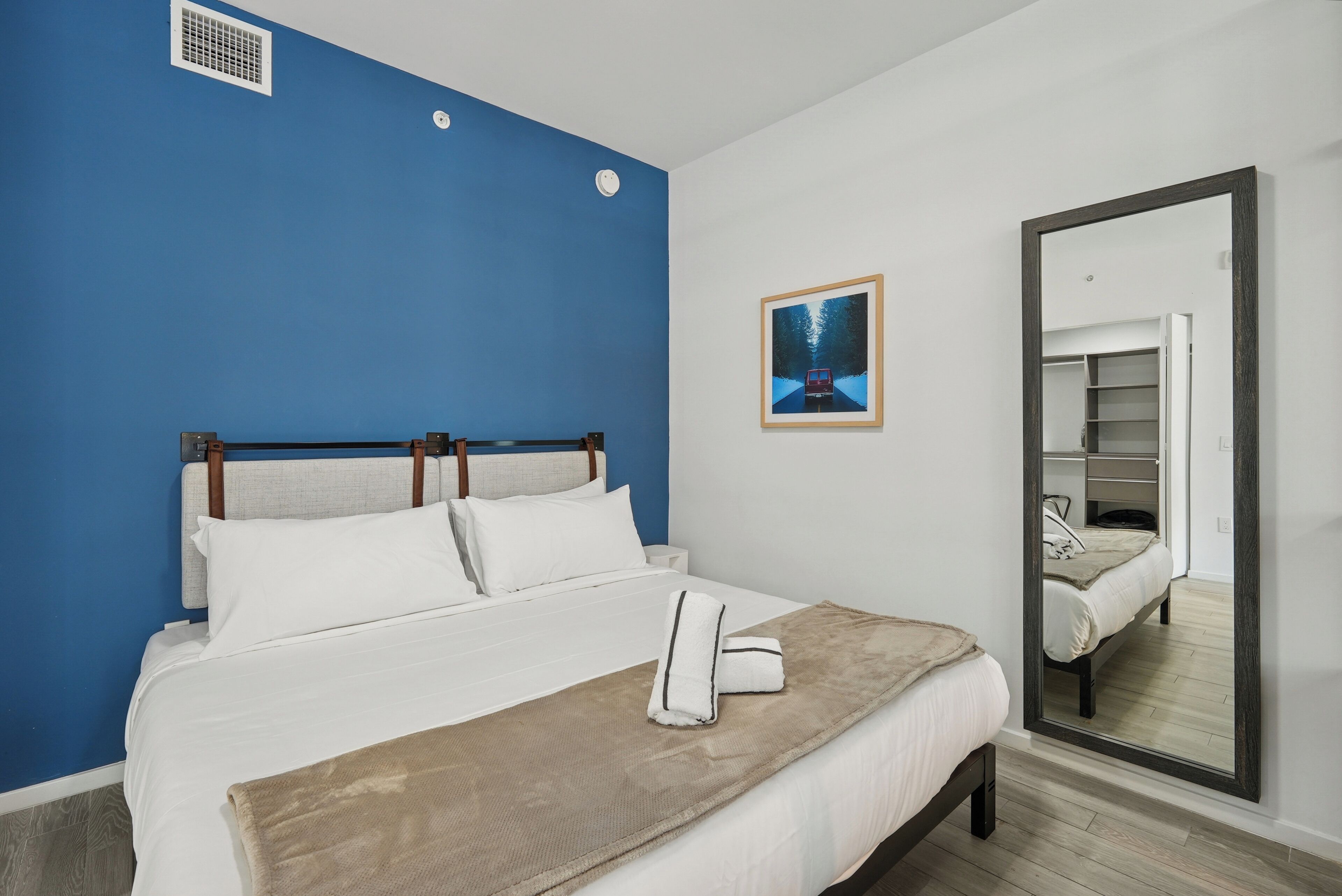 A hotel room with a large bed, a blue accent wall, a framed picture, and a mirror reflecting the room.