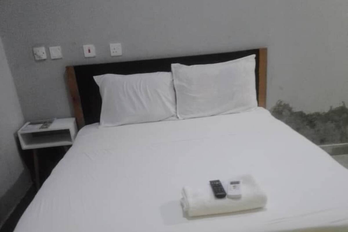 Una cama con ropa de cama blanca, un control remoto y una mesita de noche con una lámpara.