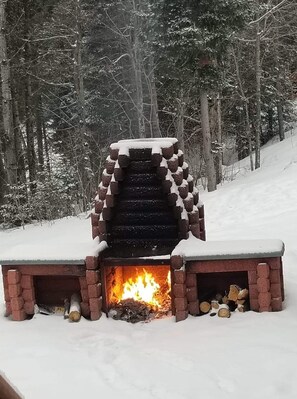 A snow-covered outdoor fireplace with a brick structure and a fire burning inside, surrounded by a snowy forest. - Bathurst New Brunswick Access to snowmobiles trail and ATV. (Saint-Laurent)