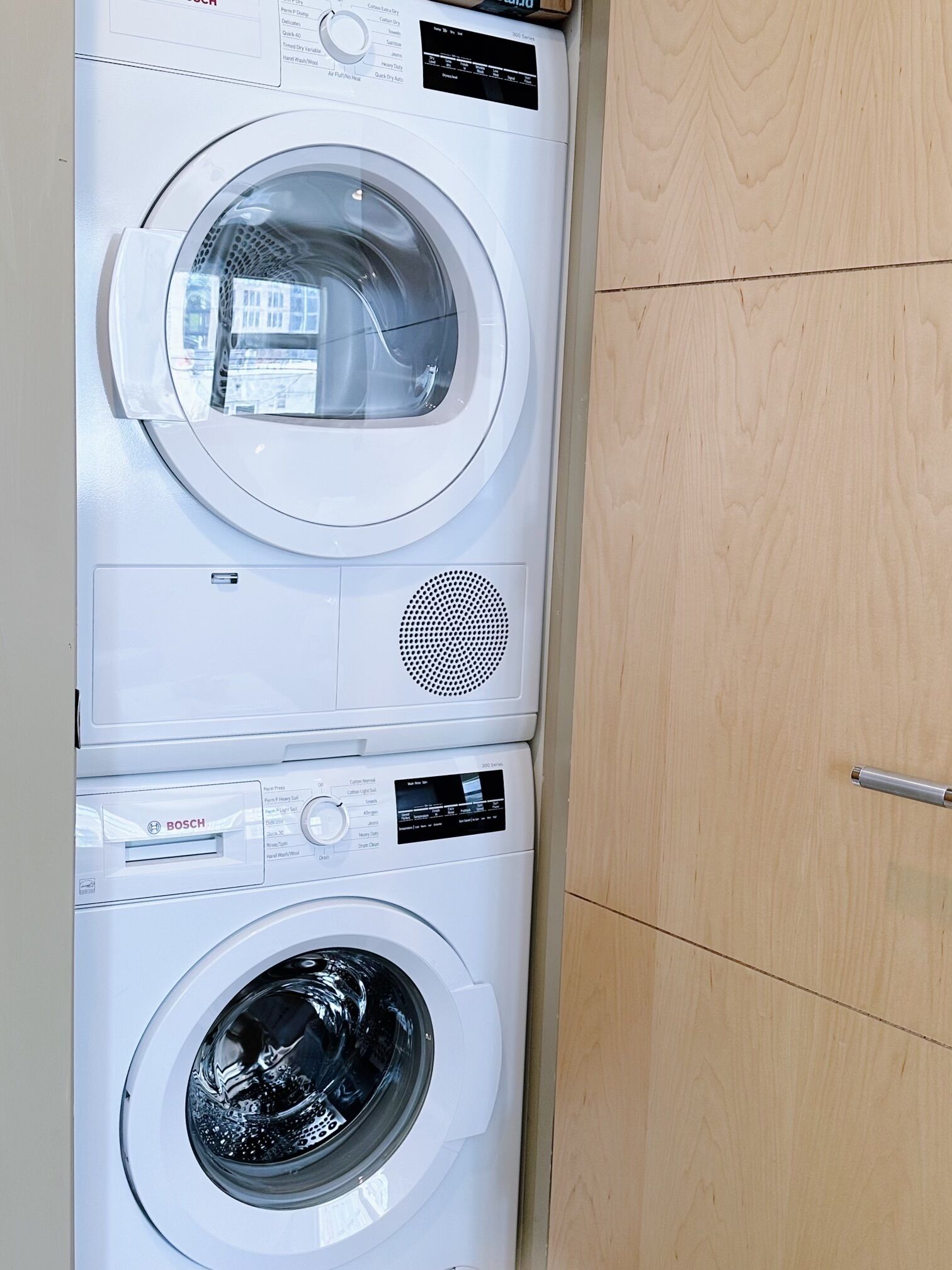 Un appareil combiné lave-linge et sèche-linge, de type « superposé », doté d’un panneau de commande et d’une porte de meuble en bois.