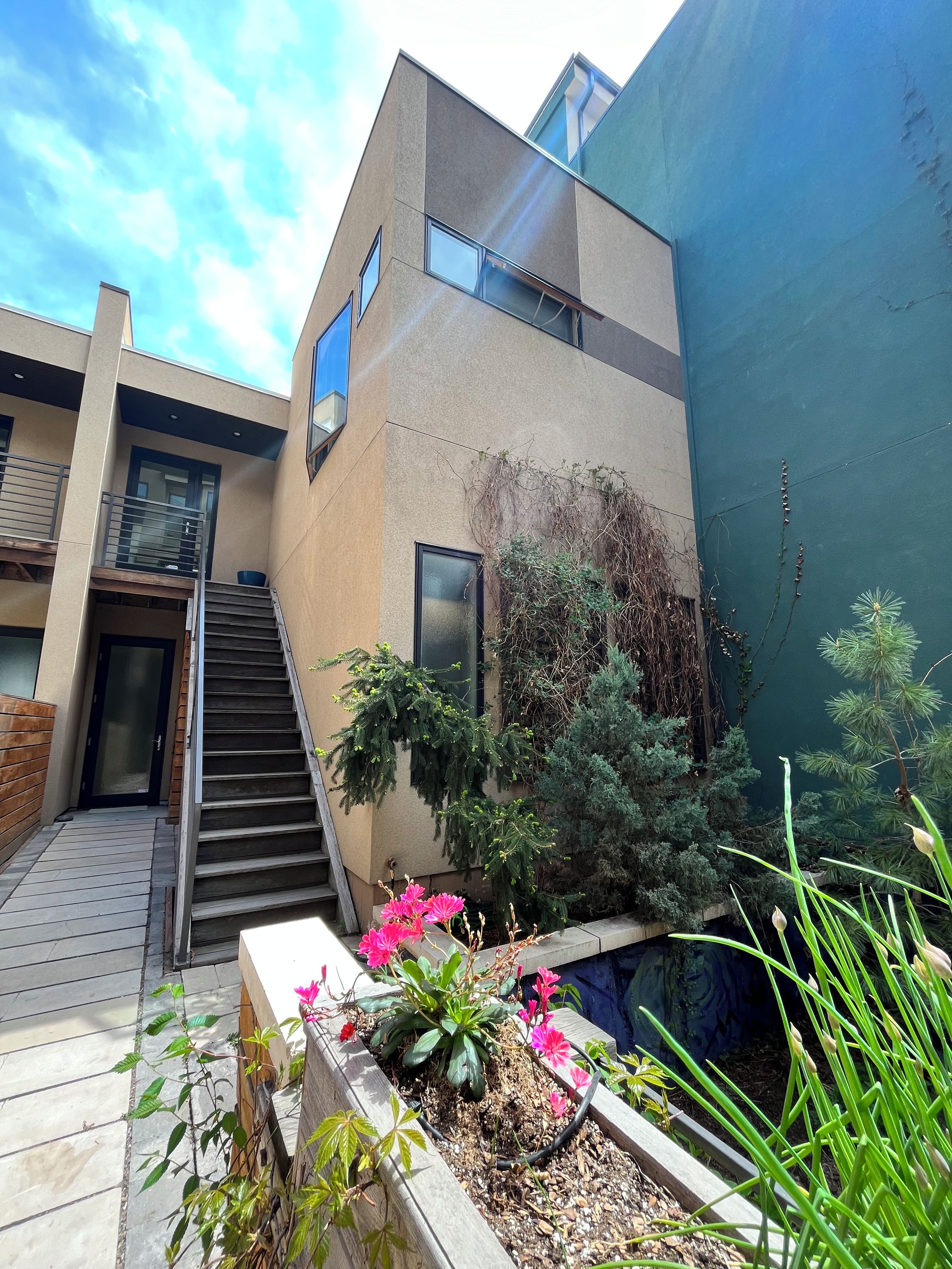 Un edificio moderno con una escalera que conduce a una entrada, un pequeño jardín con plantas y flores, y un cielo despejado.