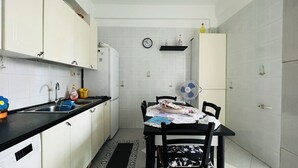 Una cocina compacta con armarios blancos, una encimera negra, una mesa de comedor con sillas, un reloj en la pared y un ventilador sobre la mesa.