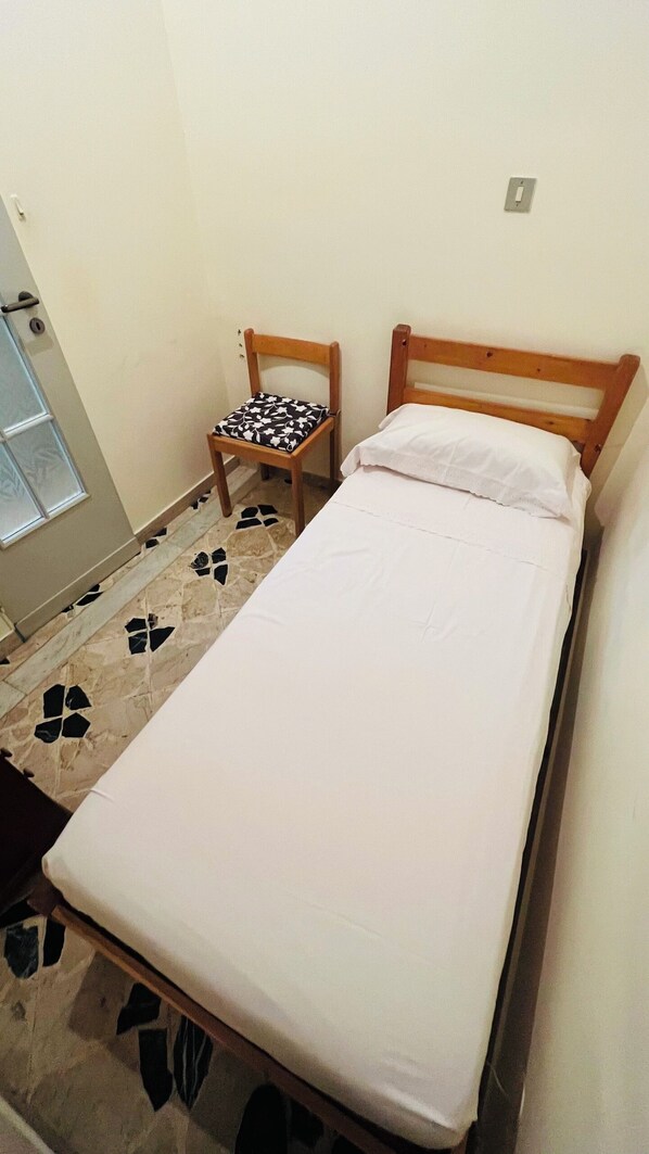 A small, single-bed room with a wooden headboard, a chair with a patterned cushion, and a tiled floor with a paw print design.