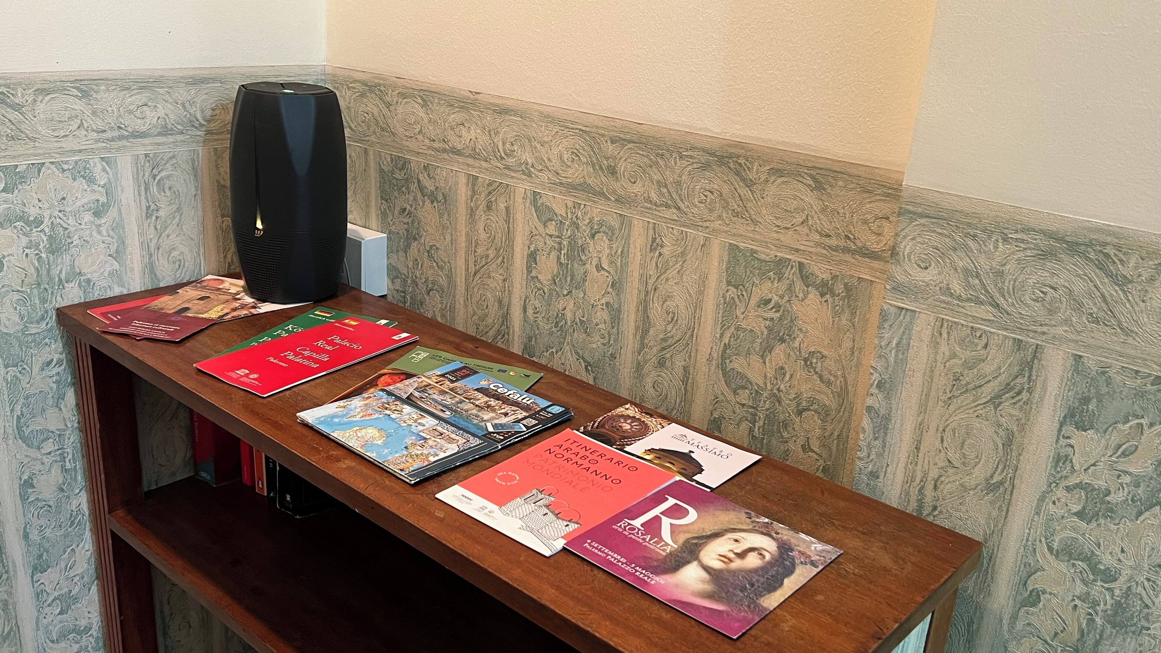 Een houten tafel met boeken en een zwarte speaker in de hoek, met behangpapier met patroon.