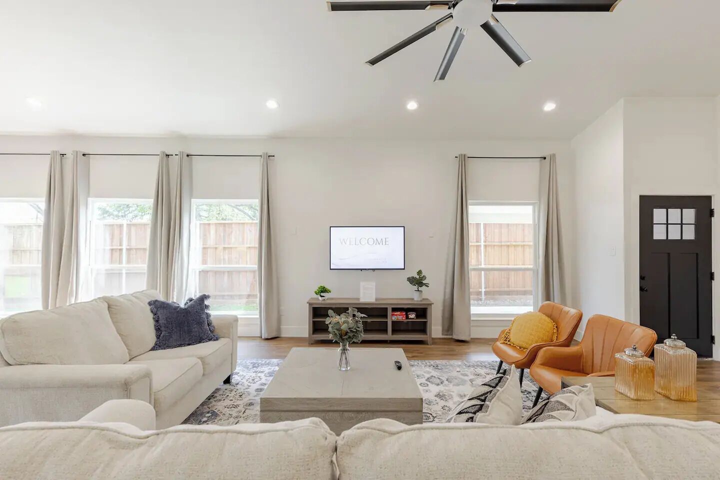 A spacious living room with a white sofa, a brown leather chair, and a coffee table. There is a TV on the wall and a door in the background.
