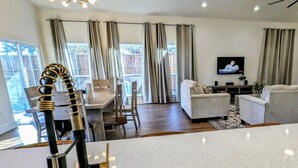 A modern kitchen with a stainless steel sink, a dining area with a wooden table and chairs, a living room with a sofa and TV, and large windows with curtains. - Spacious family/group stay near downtown (McKinney)