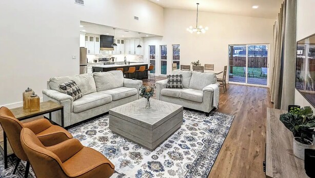 A spacious living room with a modern ceiling fan, a kitchen in the background, and a large window providing a view of the outdoors. - Spacious family/group stay near downtown (McKinney)