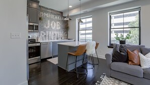 A modern kitchen with a central island, bar stools, a grey sofa with cushions, and large windows providing natural light.