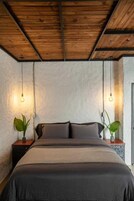 Un dormitorio con techo de madera, dos mesitas de noche con lámparas y una cama con colcha oscura.