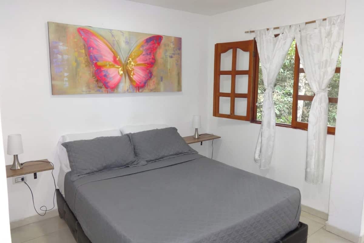 A bedroom with a bed, a nightstand, a lamp, a window with curtains, and a painting of a butterfly.